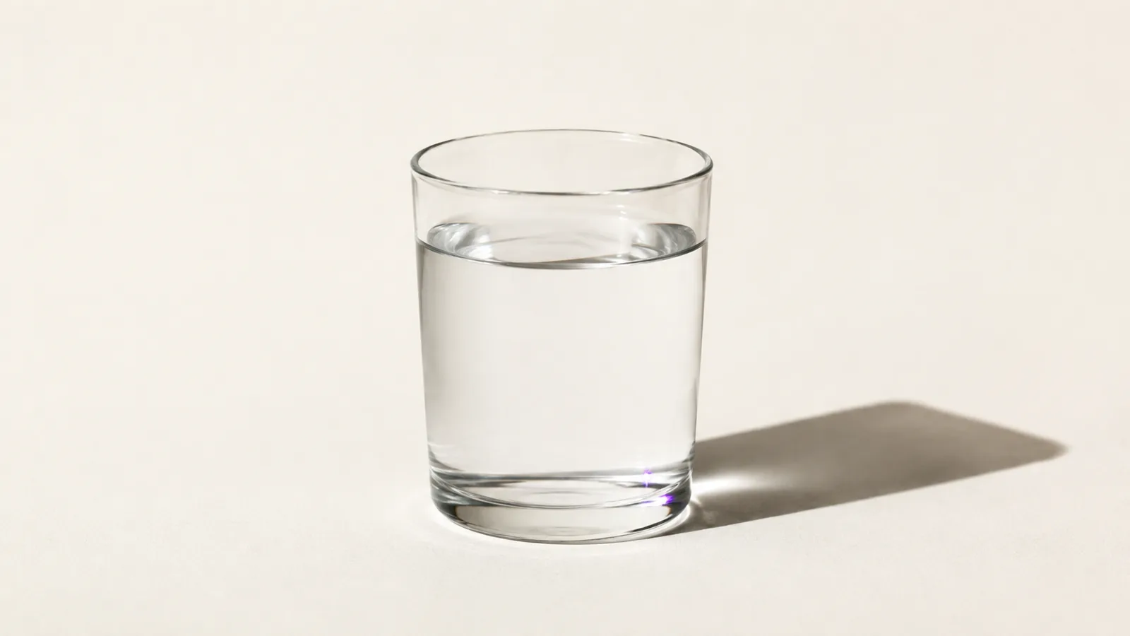 A drinking glass of clear water on a pale surface casting a sharp shadow.